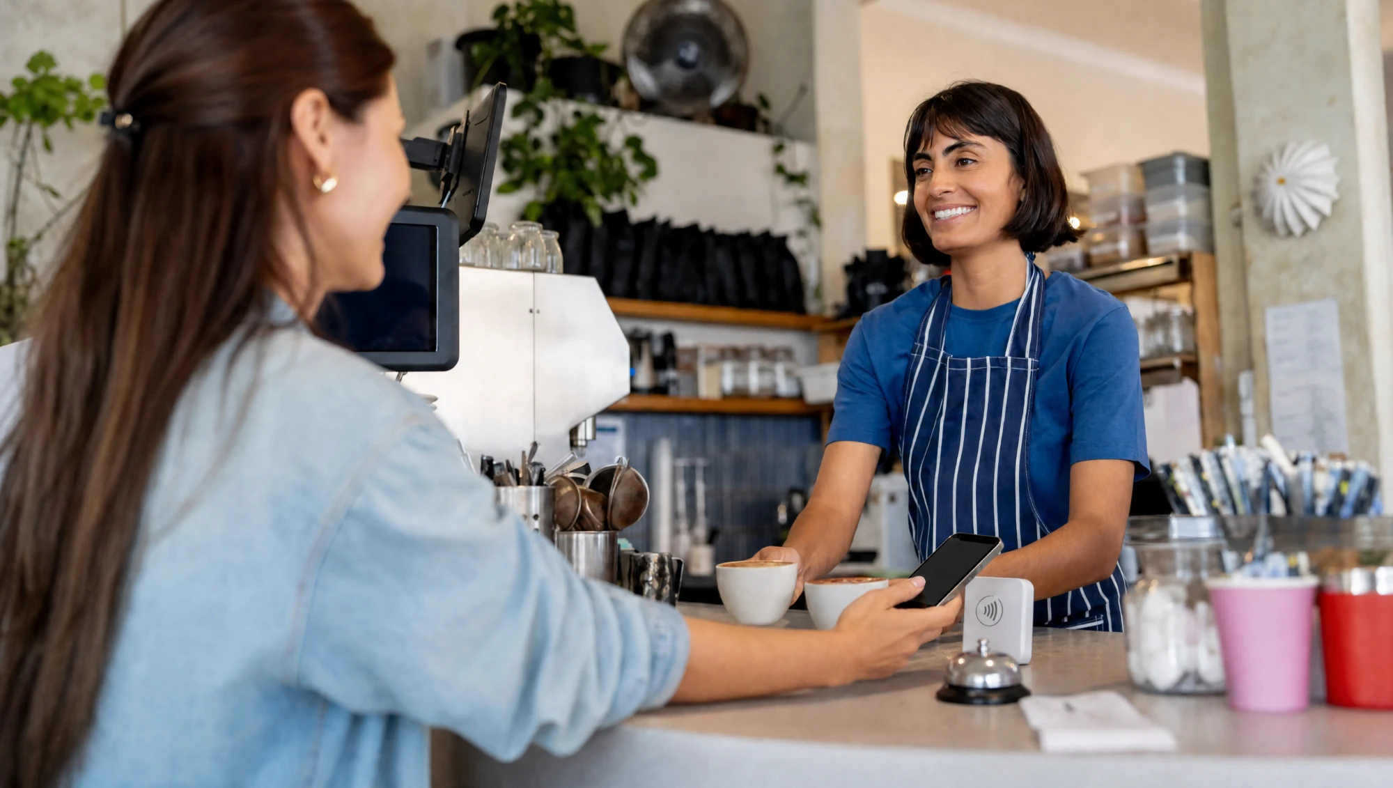 Een klant houdt haar telefoon bij het blokje op de toonbank, terwijl de barista twee koppen koffie aangeeft.